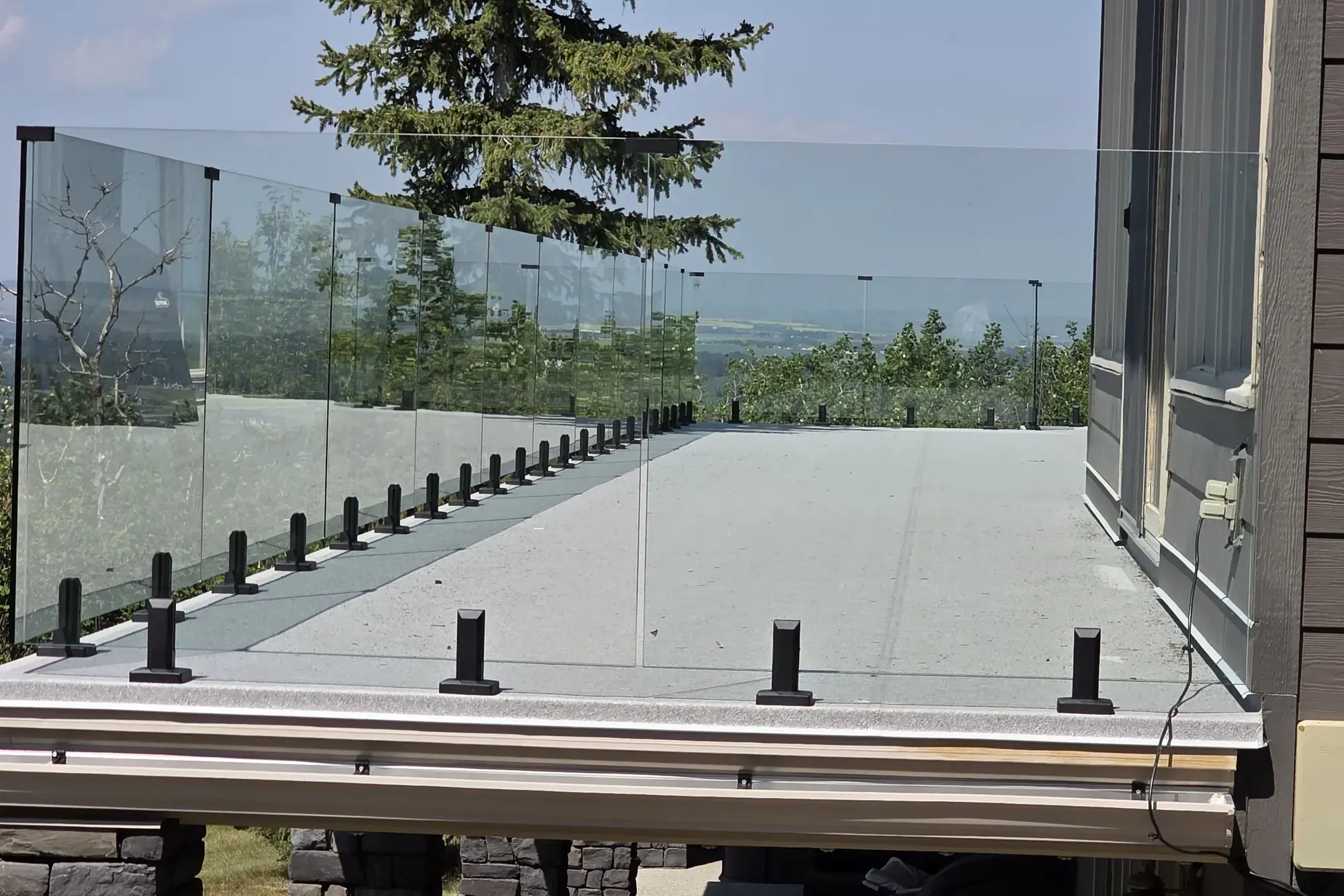 A pale granite vinyl deck beside the house behind frameless glass railing, spruce and hills beyond
