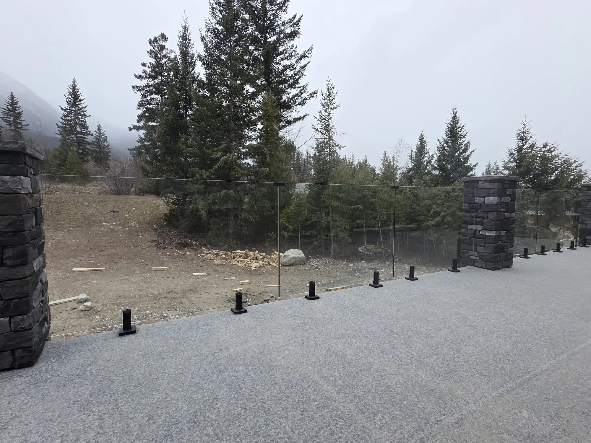 A vinyl deck between stone pillars, glass railing, misty spruce hillside beyond