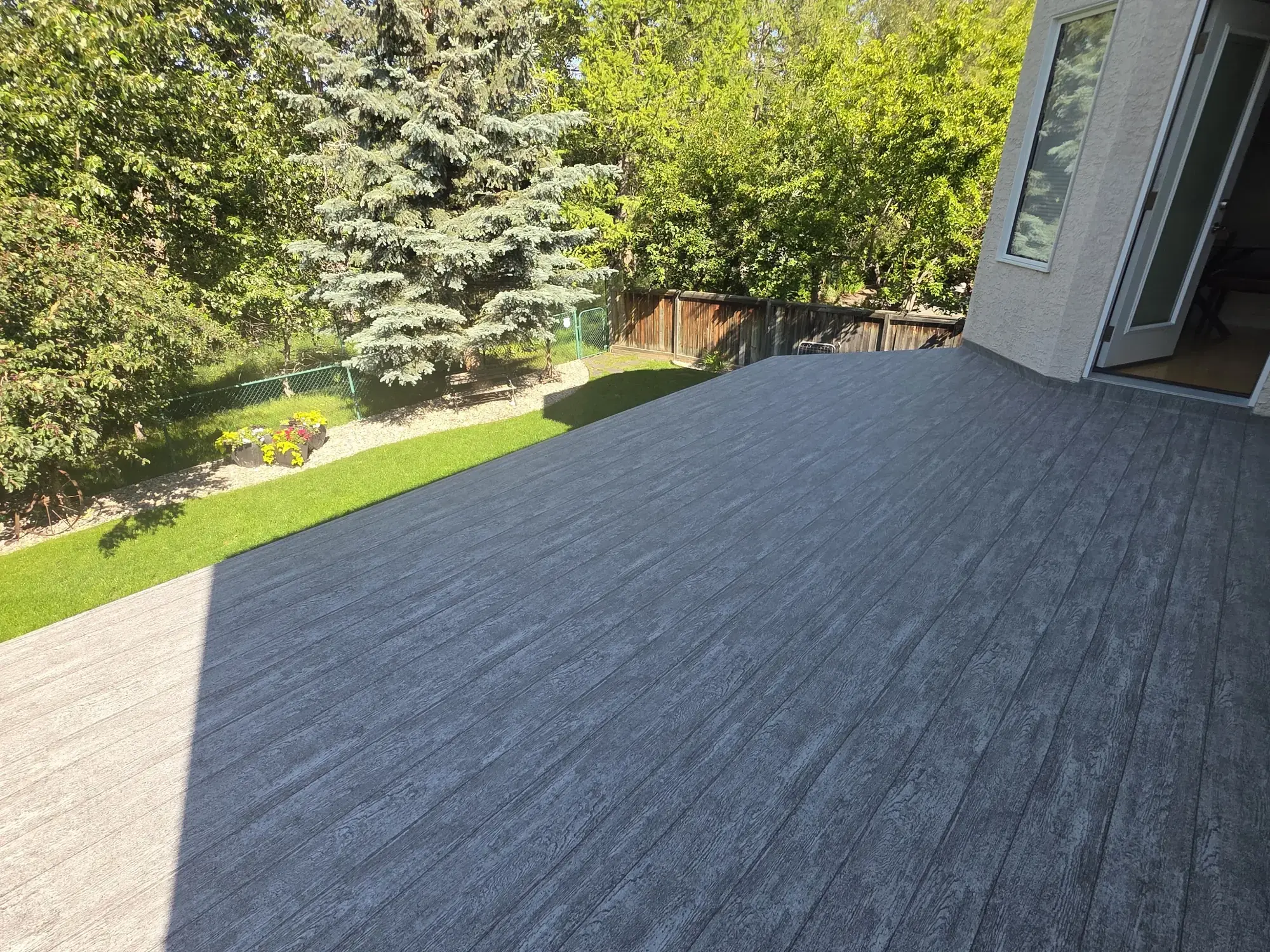 A wide woodgrain vinyl deck running from the open patio doors out to a green lawn, spruce behind