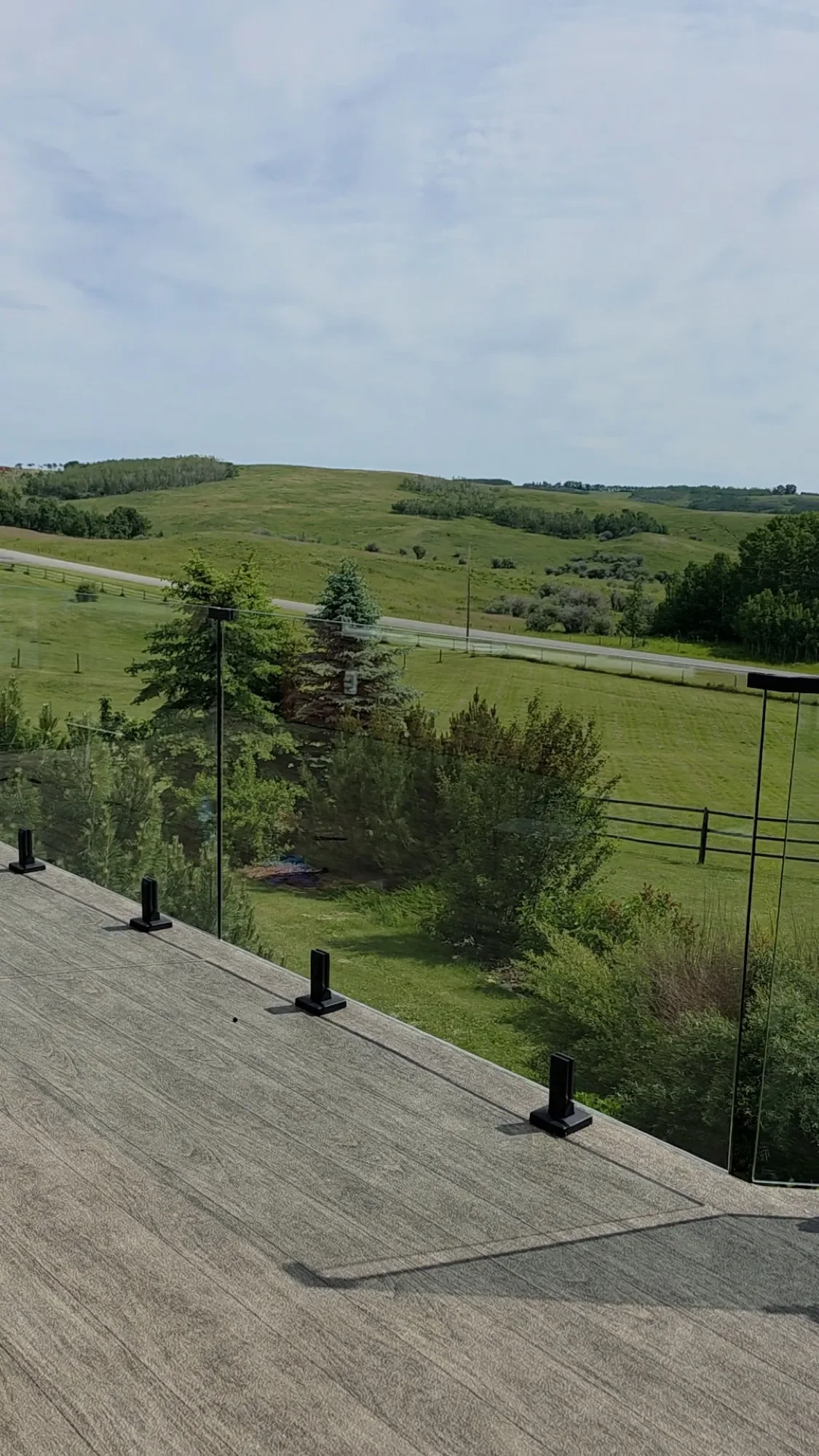 A woodgrain vinyl deck with frameless glass railing, looking out over green Alberta foothills